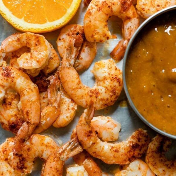 Top down view of pieces of chipotle shrimp on roasting sheet with slice of orange and bowl of mango sauce on a gray concrete surface
