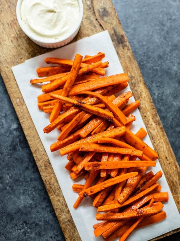 Orange harissa roasted carrots piled on a piece of parchment on a brown cutting board with bowl of garlic lemon aioli all on gray concrete surface