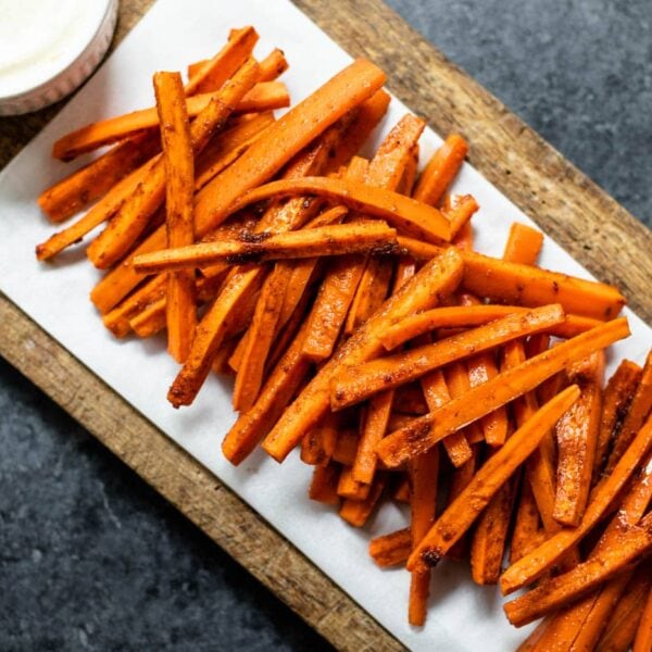 Orange harissa roasted carrots piled on a piece of parchment on a brown cutting board with bowl of garlic lemon aioli all on gray concrete surface