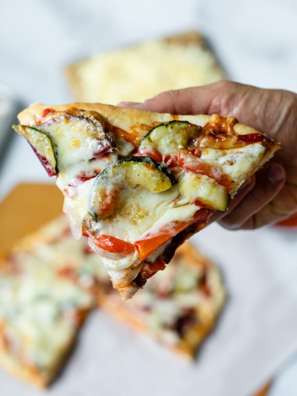 Piece of homemade roasted vegetable pizza being held with rest of pizza in background on top of white parchment with pizza board and extra cheese on butting board all on marble surface