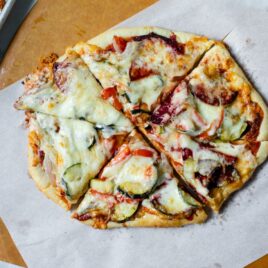 Top down view of sliced of roasted vegetable pizza on top of white parchment paper on pizza board with extra bowl of sauce and pan of roasted vegetables on marble surface