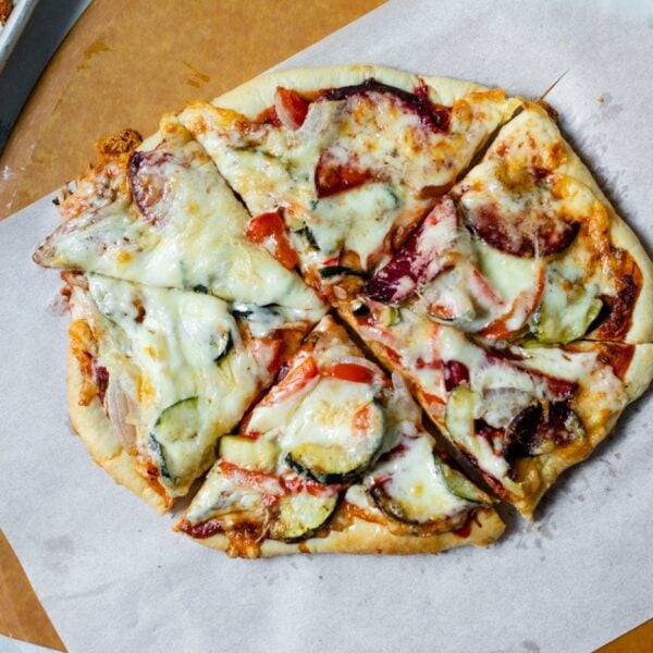 Top down view of sliced of roasted vegetable pizza on top of white parchment paper on pizza board with extra bowl of sauce and pan of roasted vegetables on marble surface