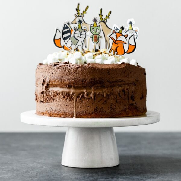 A s'mores ice cream cake covered in chocolate frosting, topped with torched marshmallows and birthday animal figurines on a white cake plate on a gray slate surface with white background