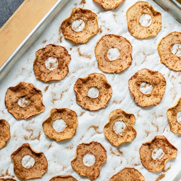 Top down view of homemade apple chips with fall spices on top sitting on a parchment-lined baking sheet on a wood board with apples to the side on a gray slate surface