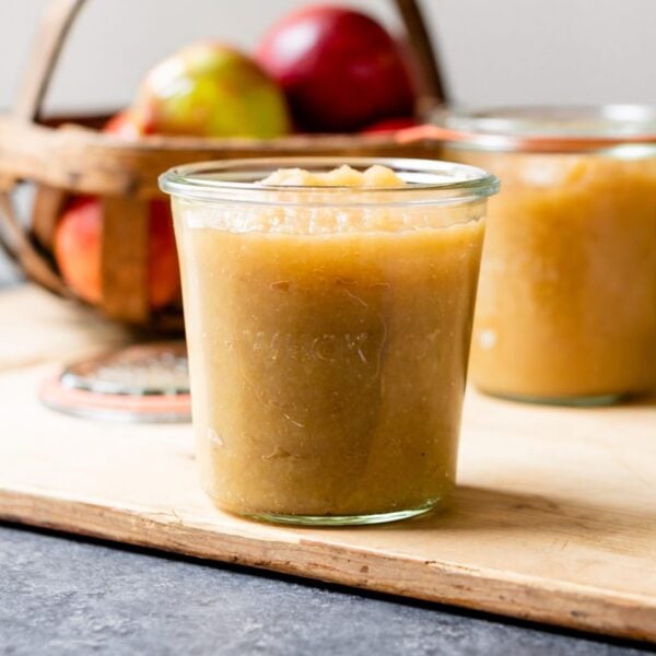 Glass jar of homemade applesauce sitting on wood board with basket of apples and two glass jars in background on gray slate surface