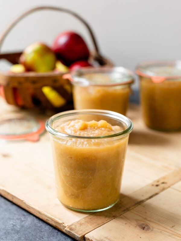 Glass jar of homemade applesauce sitting on wood board with basket of apples and two glass jars in background on gray slate surface
