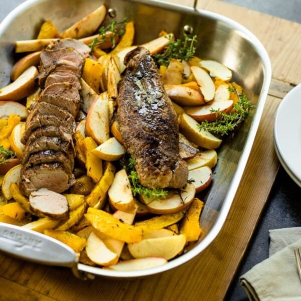 Roasted pork tenderloin sitting on bed of apples and squash in a roasting pan on a wood board with white plate and silverware on a gray slate surface