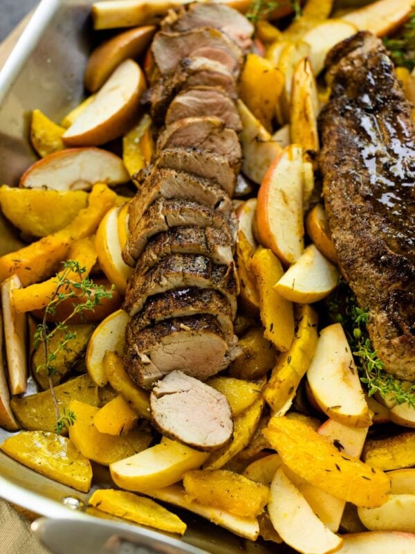 Roasted pork tenderloin sitting on bed of roasted apples and squash in a roasting pan on wood board with gray slate surface in background