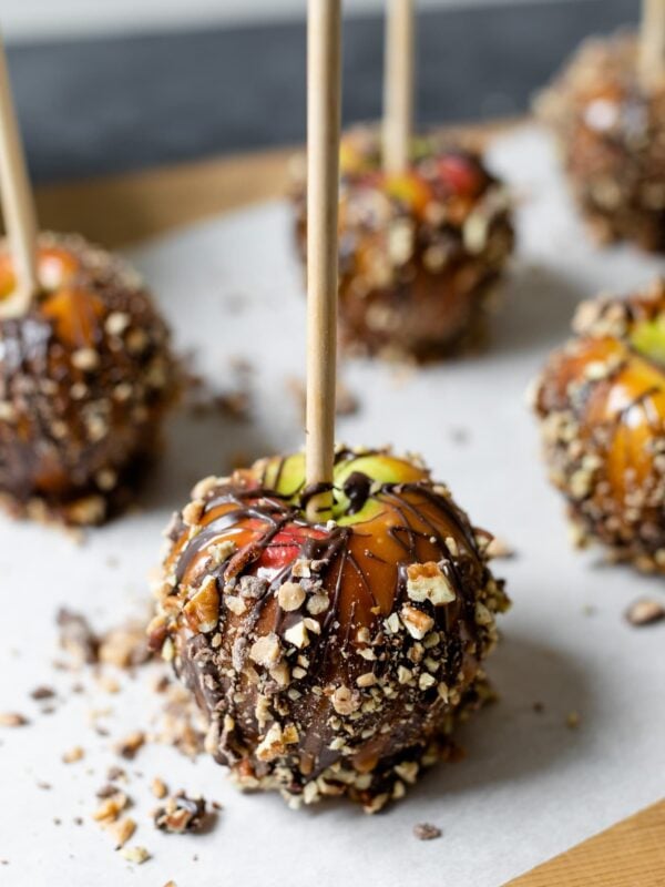 Homemade caramel apples sitting on piece of parchment sprinkled with toffee pieces and pecan pieces on a wood board with gray slate background