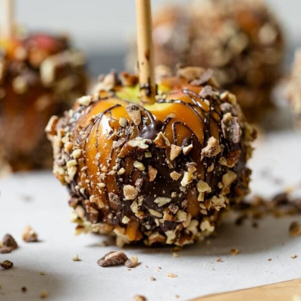 Homemade caramel apples sitting on parchment-lined baking sheet sprinkled with toffee pieces and chopped pecans with chocolate all on wood board with white background