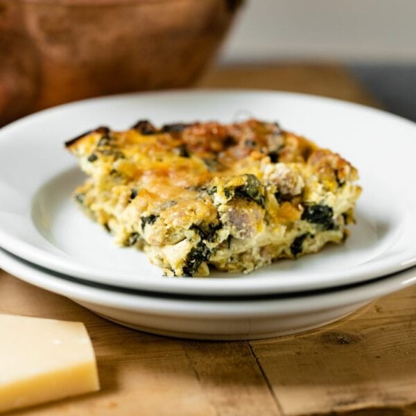Piece of squash kale lasagna sitting on white plate with extra white plate underneath with piece of Parmesan cheese on wood board with copper pot in background all on gray slate surface
