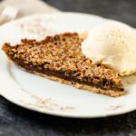 Pecan pie bars sitting on plate with scoop of vanilla ice cream with linen napkin and fork all on gray slate surface