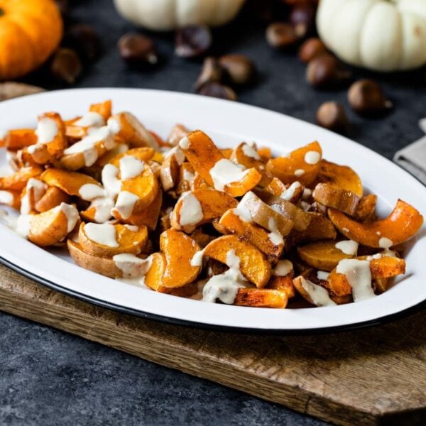 Spiced butternut squash sprinkled with tahini sauce in a white platter sitting on wood board with pumpkins and nuts in background on gray slate surface