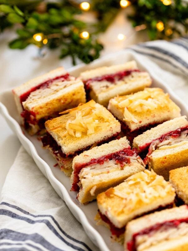 Turkey cranberry croque monsieur sandwiches sitting on white serving plate with linen napkin underneath with Christmas lights and greenery in background on white surface