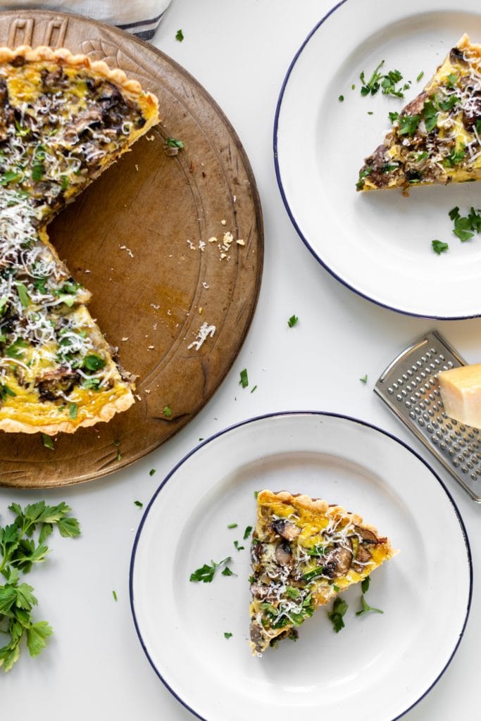 Breakfast quiche topped with parsley and Parmesan cheese with pieces cut out on bread board with extra cheese and parsley all sitting on white surface