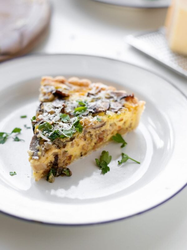 Breakfast quiche topped with parsley and Parmesan cheese sitting on white plate with extra cheese and bread board in background all on white surface