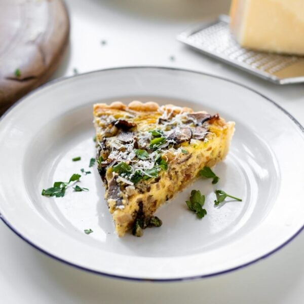 Breakfast quiche topped with parsley and Parmesan cheese sitting on white plate with extra cheese and bread board in background all on white surface