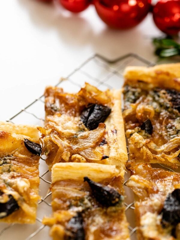 Caramelized onion tart dotted with figs and blue cheese sitting on cooling rack with red Christmas ornaments in background all on white surface