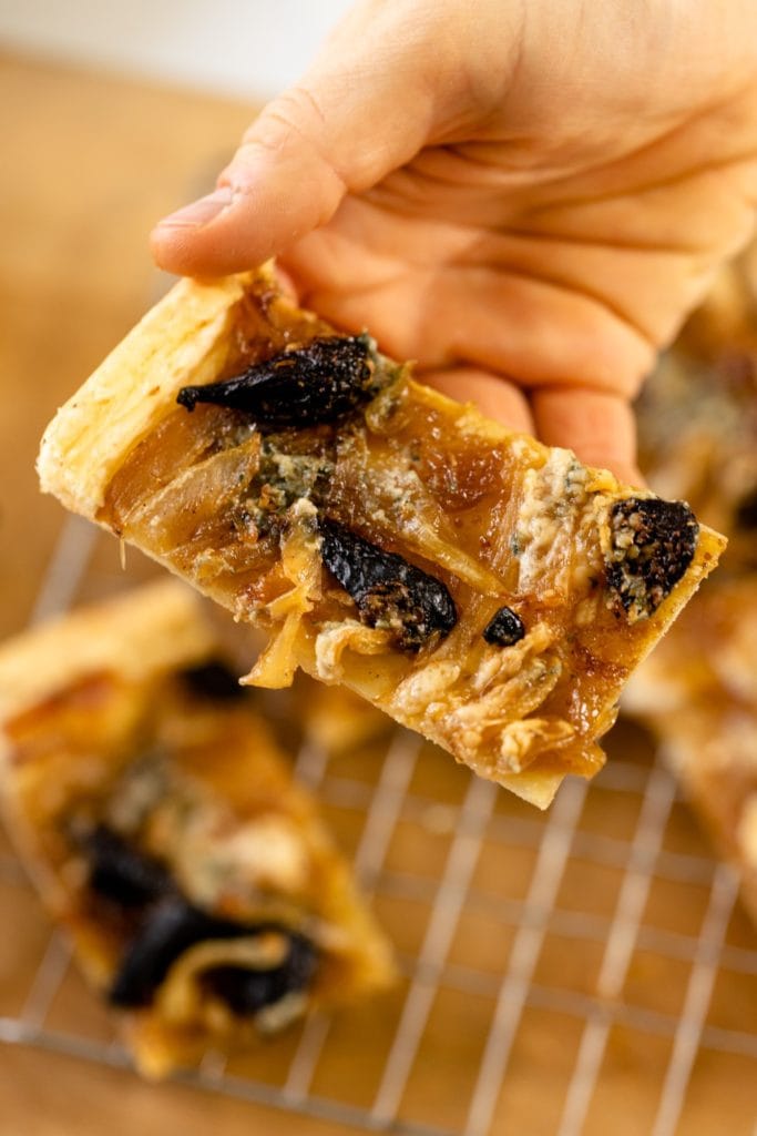Caramelized onion tart topped with dried figs and blue cheese being held by hand with extra tarts on cooling rack on wood surface in background
