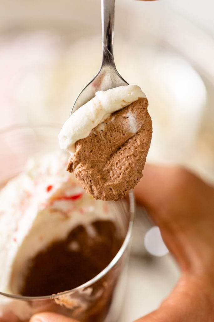 Chocolate mousse sitting on spoon held by hand with extra glasses of chocolate mousse topped with whipped cream and peppermint bits in background