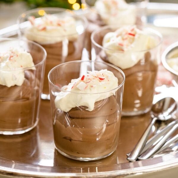 Glasses filled with chocolate mousse and topped with whipped cream and crushed peppermint pieces sitting on silver tray with container filled with whipped cream