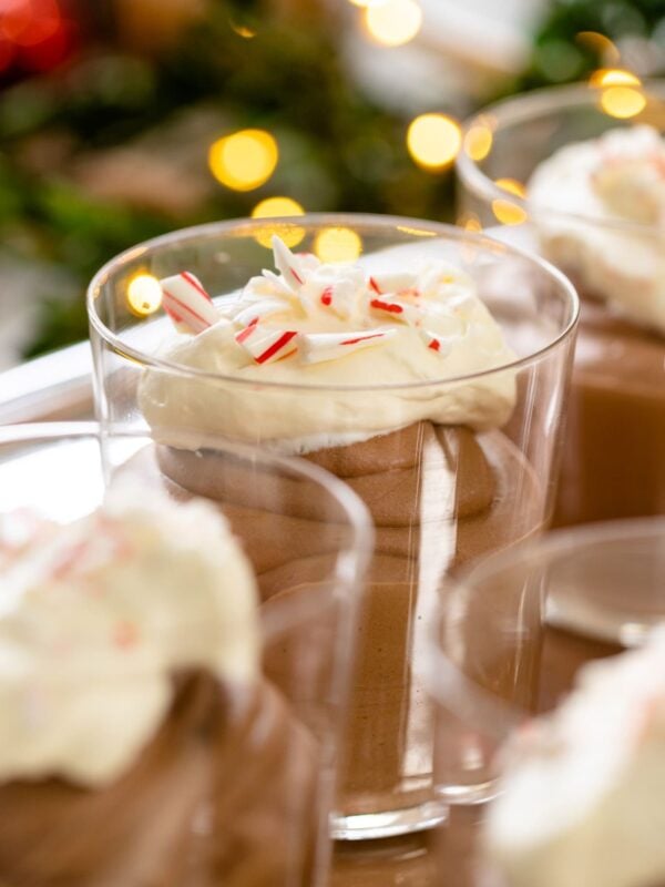 Glass filled with chocolate mousse topped with whipped cream and peppermint pieces with other glasses filled with mousse and Christmas lights in background