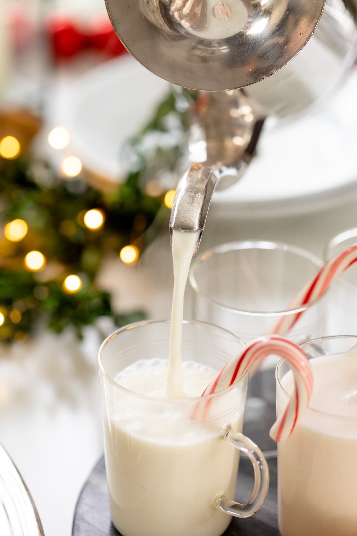 Stainless steel kettle pouring white hot chocolate into glasses with candy cane sitting on edge all on white surface with Christmas lights in background