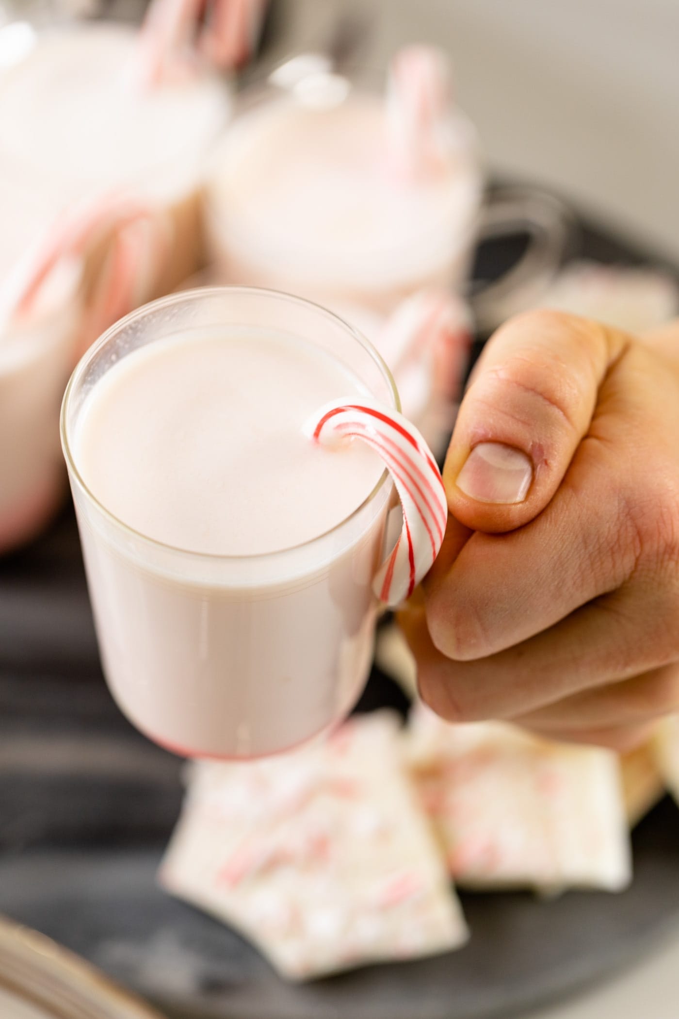 Hand holding glass of peppermint-flavored white hot chocolate with candy cane sitting on edge of glass with extra servings and peppermint bark in background