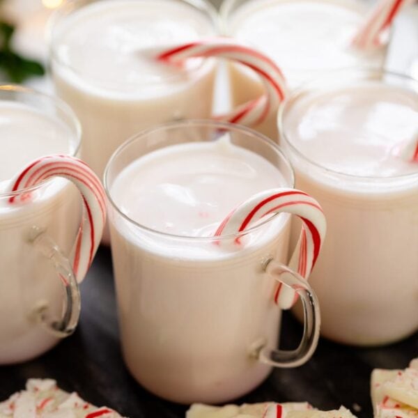 White hot chocolate in glasses with candy cane inside with extra pieces of peppermint bark all on white surface