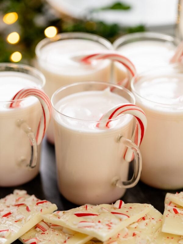 Peppermint flavored white hot chocolate in glass mug with candy cane sitting on edge with extra peppermint bark in front all on white surface with Christmas lights in background