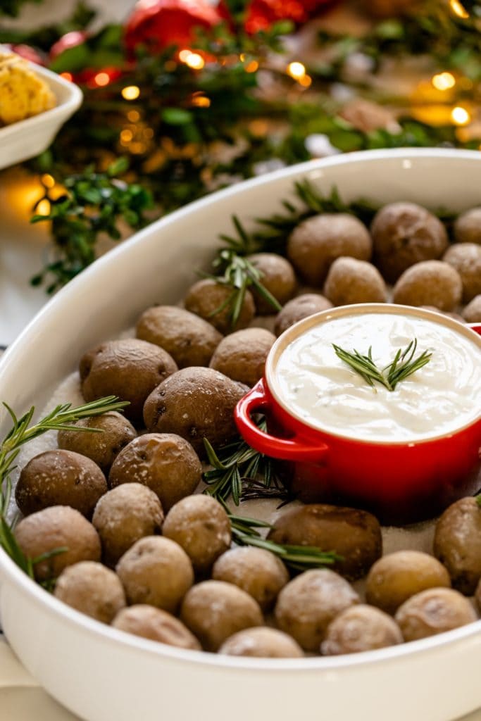 Salt roasted potatoes sitting on bed of salt in white roasting dish with rosemary ranch dipping sauce in red container with Christmas lights in background