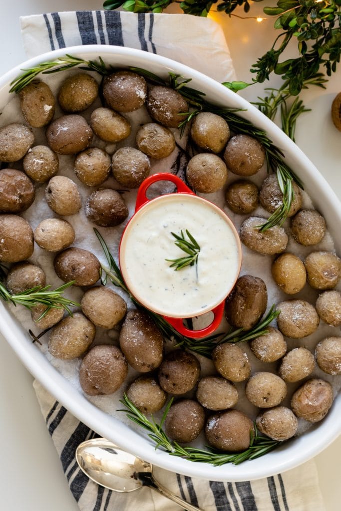 Salt roasted potatoes sitting on bed of salt in white roasting dish with rosemary ranch sauce in red container placed in center with Christmas lights all on white surface