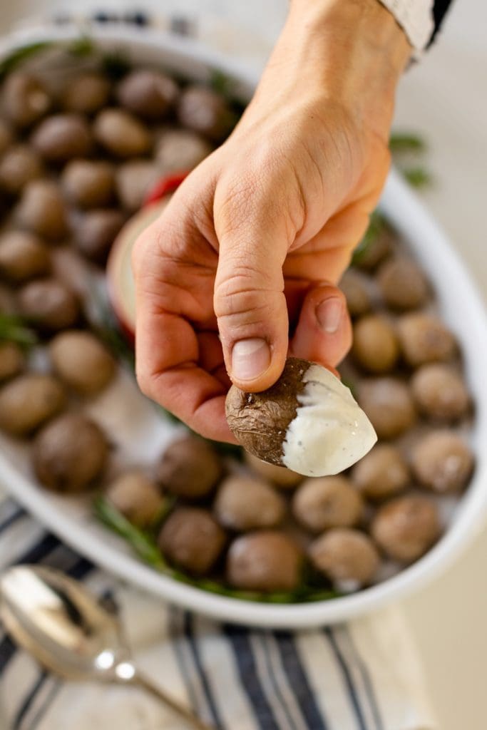 Salt roasted potatoes being held by hand with rosemary ranch sauce on end of potatoes all on white surface with striped blue linen and serving spoon
