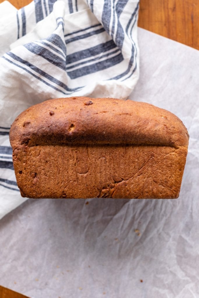 Side view of whole loaf of hearty grain bread with towel and parchment in background all on wood surface