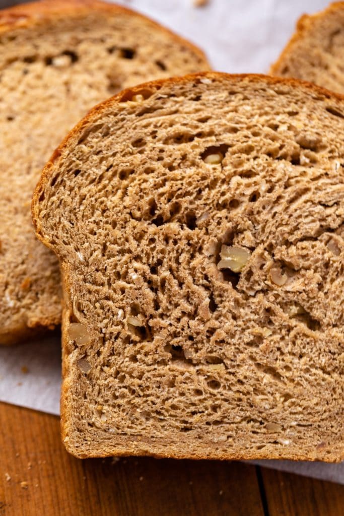 Close up view of inside of one piece of hearty grain bread with extra slices of bread in background on wood surface with parchment underneat