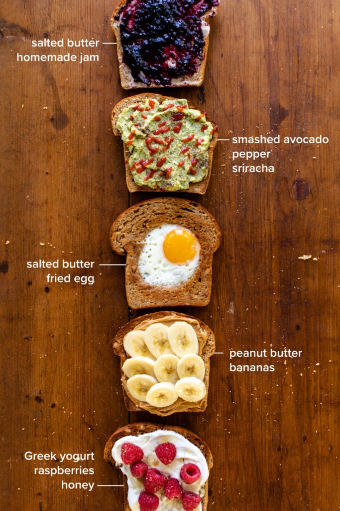 Five versions of hearty grain bread sitting on piece of wood, each topped with a unique combination including jam, avocado, an egg, banana and peanut butter and yogurt with raspberries