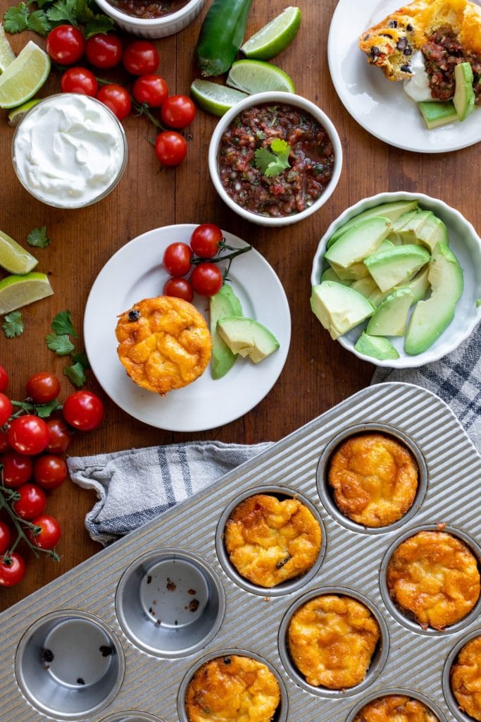 Huevos rancheros egg cups sitting on white plates with pan filled with extra cups as well as many toppings including salsa, sour cream and guacamole all on wood surface