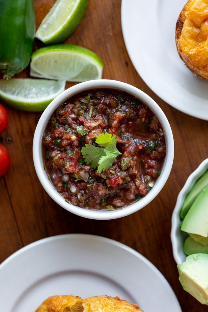 Homemade restaurant-style salsa sitting in white bowl with cilantro leaf on top surrounded by lime wedges and avocado all on wood surface
