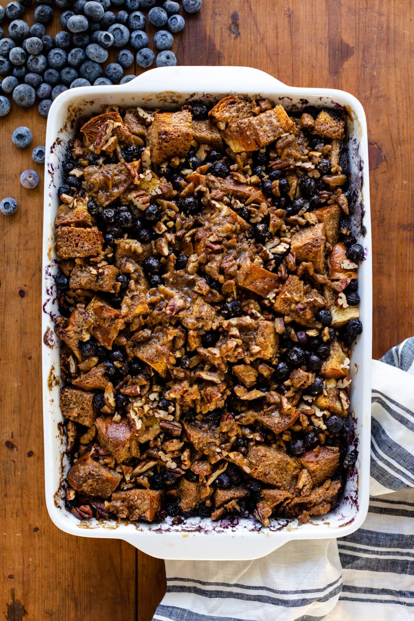 Top down view of overnight blueberry French toast in white baking dish with striped white towel and extra blueberries all on wood surface