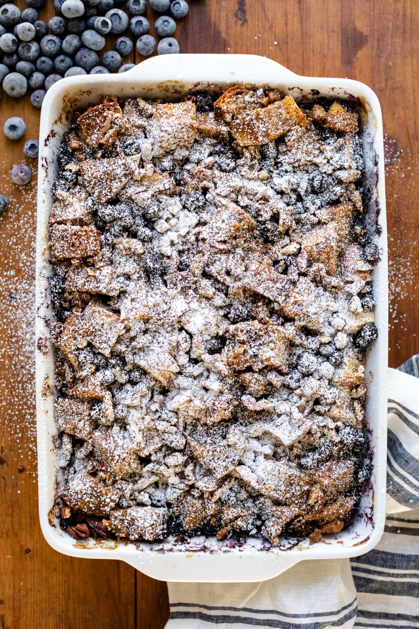 Top down view of overnight blueberry French toast sitting in a white bowl topped with powdered sugar with blueberries and linen towel to the sides all on a wood surface
