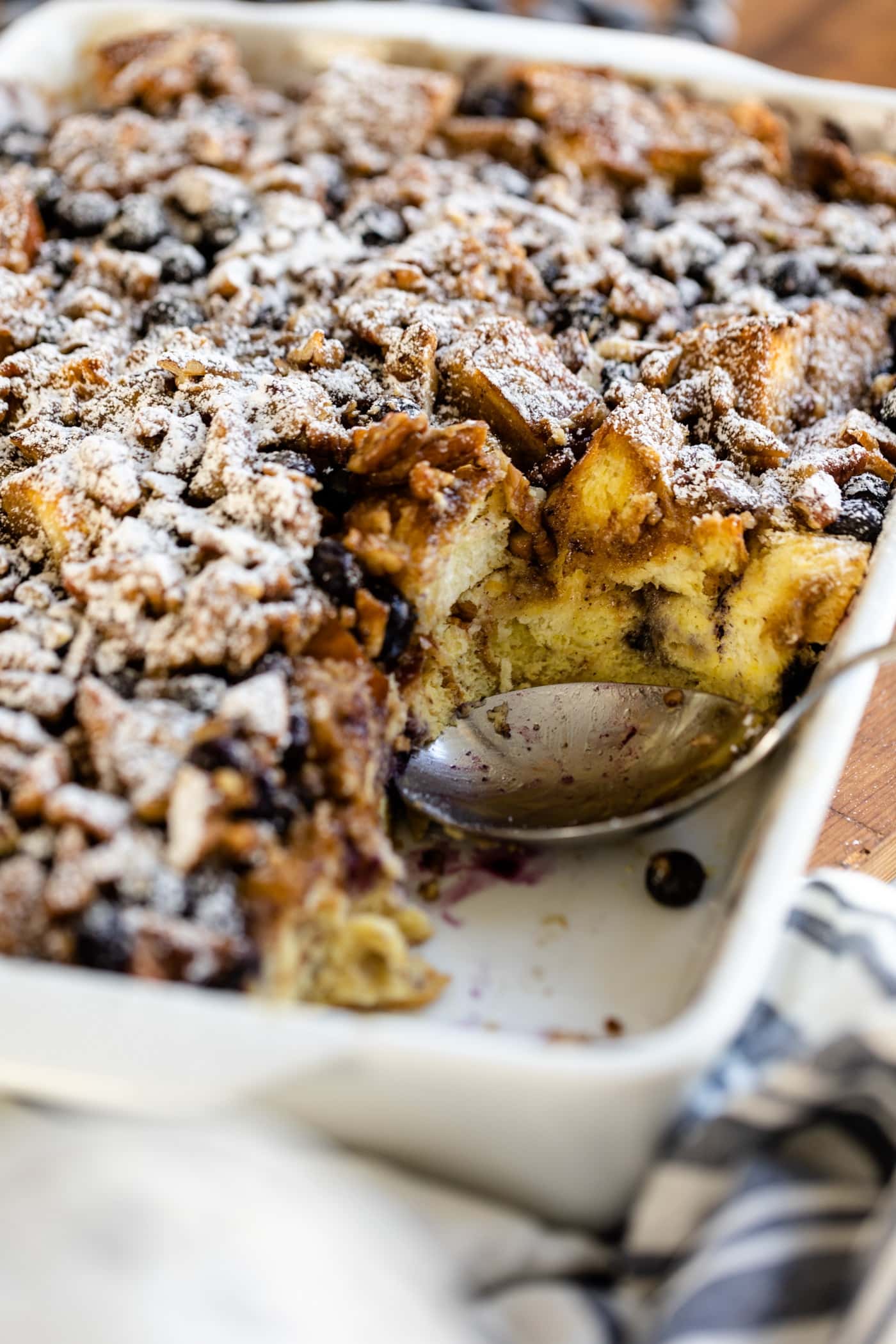 Spoon sitting in white pan filled with overnight blueberry French toast with towel in foreground and extra blueberries in background