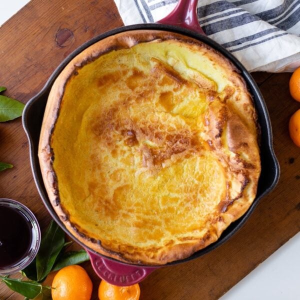 Top down view of citrus Dutch baby sitting on wood board surrounded by oranges and berry coulis in container all on white surface