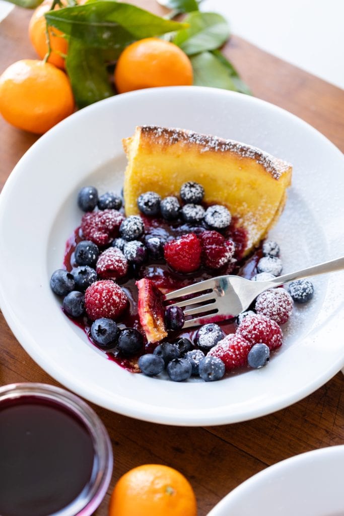 A piece of citrus Dutch baby pancake on white plate topped with berries and powdered sugar with fork sitting on edge all on wood surface surrounded by oranges