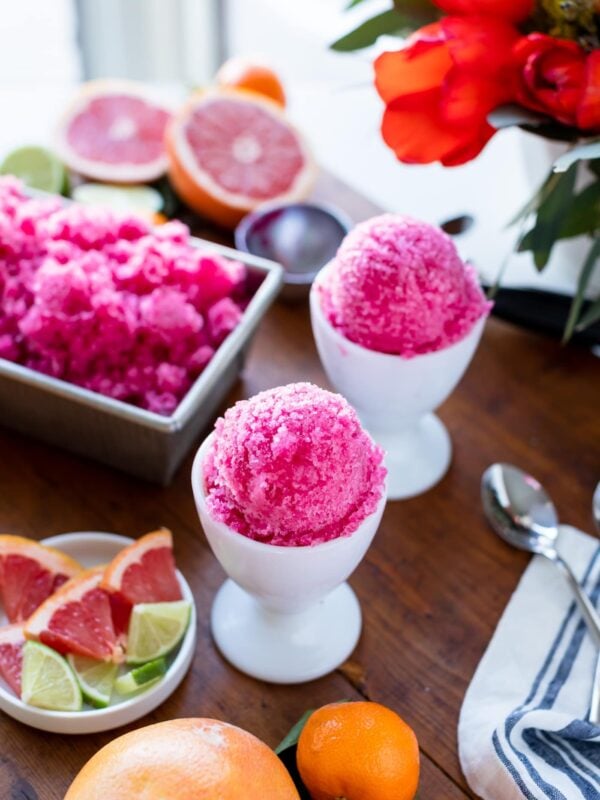 Two white cups holding grapefruit flavored fruit ice surrounded by fresh fruit and extra servings of fruit ice all on wood board with flowers in background