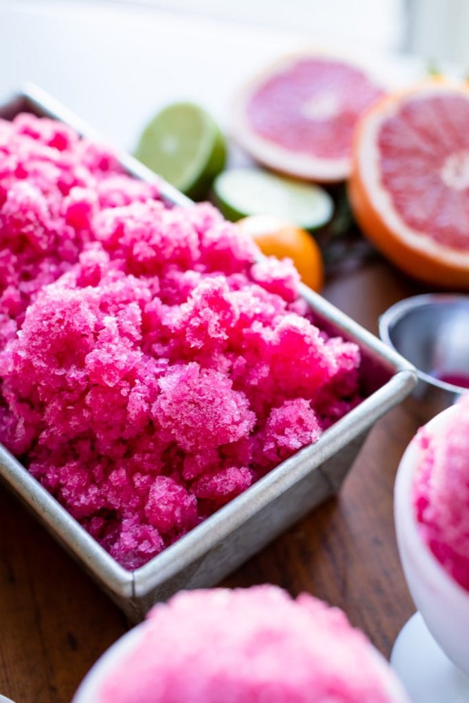 Stainless steel loaf pan filled with grapefruit-flavored pink fruit ice with extra fruit in background all on wood surface