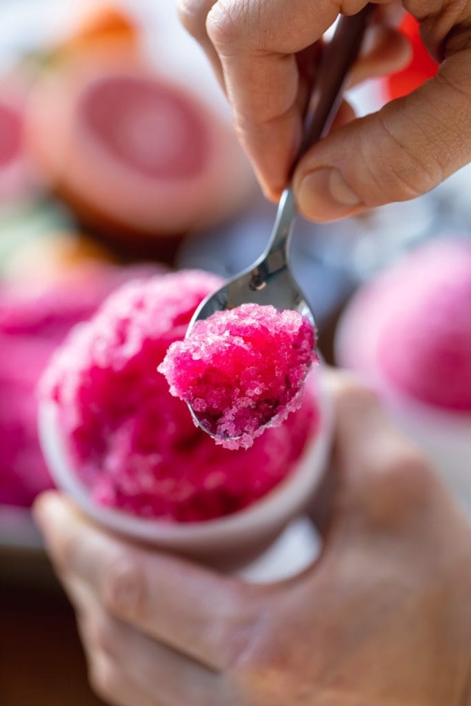 Hand holding spoonful of pink frozen ice with extra fruit ice in background in white cup
