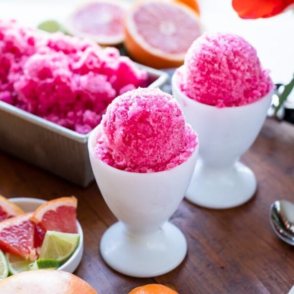 Two white cups holding grapefruit flavored fruit ice surrounded by fresh fruit and extra servings of fruit ice all on wood board