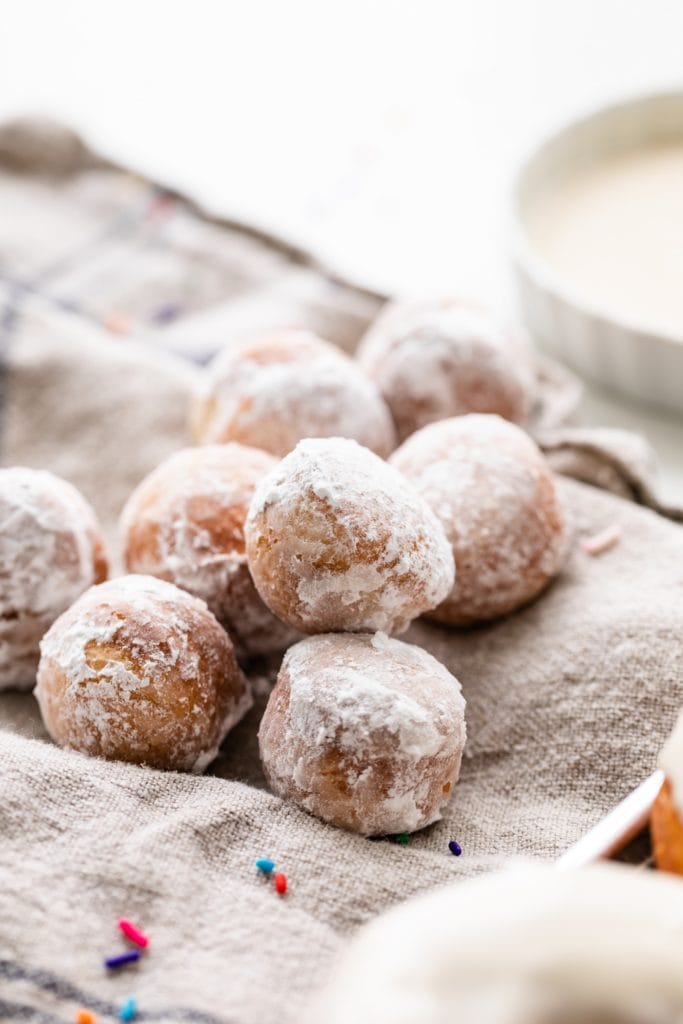 Pile of homemade donut holes topped with powdered sugar sitting on linen towel
