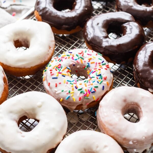 Homemade yeast donuts covered in vanilla and chocolate glazes sitting on wire cooling rack with linen towel and extra donut holes in background