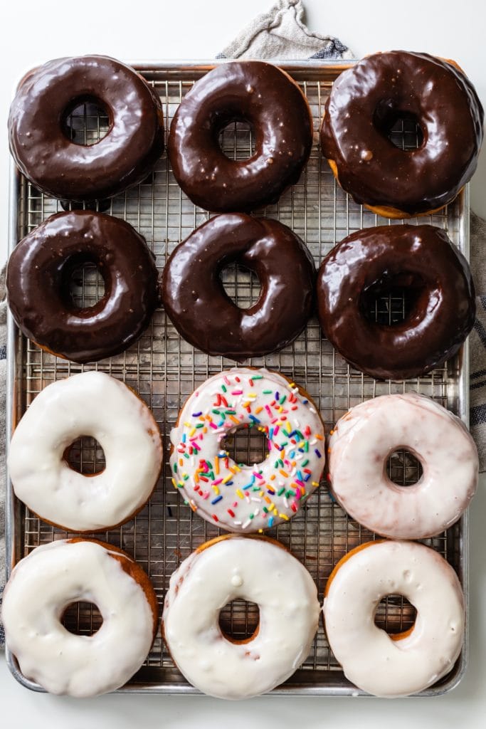 Top down view of a dozen homemade yeast donuts sitting on wire cooling rack with towel underneath all on a white surface